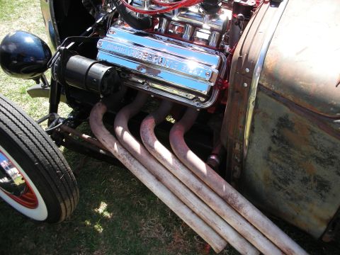 1998 Ford Crown Victoria - Overhead view of a chrome-finned V8 with rusted swept zoomie exhaust headers and a whitewall tire in a bare-metal hot rod.