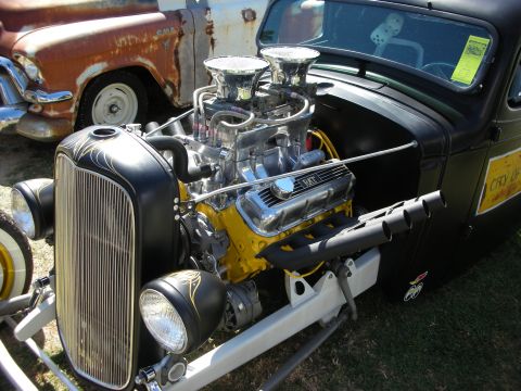 1998 Ford Crown Victoria - black hot rod pickup with a pinstriped oval grille and chrome dual-carb engine topped by twin velocity stacks, parked near a rusty truck.