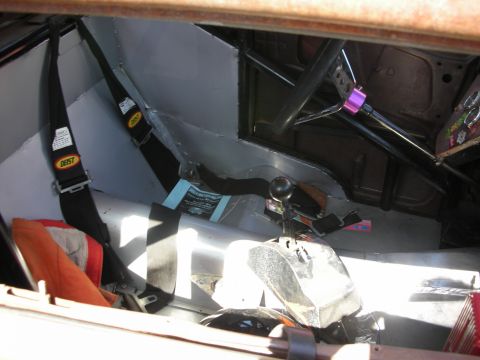 1998 Ford Crown Victoria - Bare-metal stripped interior of a race-style car showing a floor shifter, black safety harnesses and an orange bucket seat.