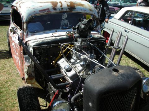 1998 Ford Crown Victoria - weathered rat rod coupe with exposed chrome V8, upright zoomie pipes and a mounted animal skull ornament, parked at a car show.