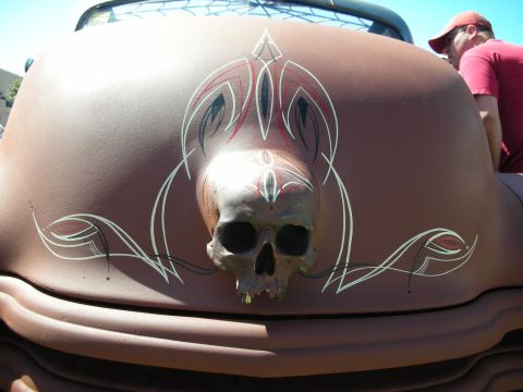 1998 Ford Crown Victoria - skull mounted on a pink-primer hood surrounded by symmetrical silver and red pinstriped scrollwork, with a person at the side.