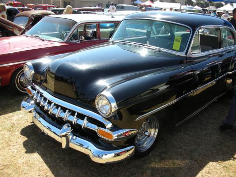 1998 Ford Crown Victoria - Glossy black early-1950s Chevrolet sedan with toothy chrome grille and steel wheels parked on grass at a car show.