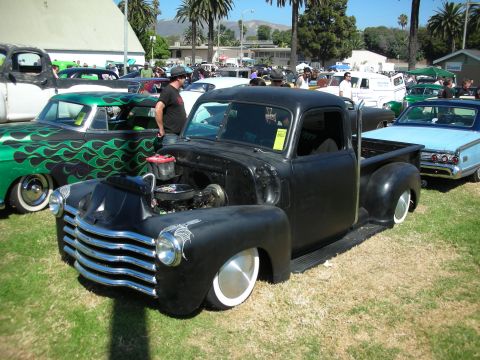 1998 Ford Crown Victoria - Lowered matte-black 1940s-50s Chevrolet pickup with exposed engine and chrome grille on a crowded car-show field.