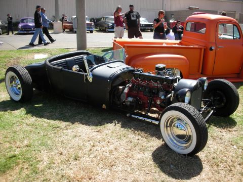 1998 Ford Crown Victoria - Flat-black channeled roadster hot rod with exposed flathead-style V8 and whitewall tires parked beside an orange pickup.