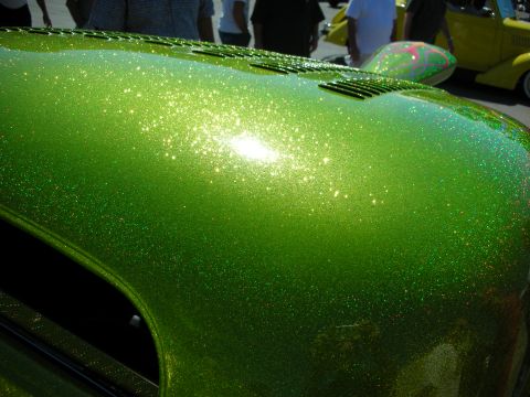 1998 Ford Crown Victoria - Detail of a curved body panel coated in vivid green metalflake paint glittering in the sun, with show-goers and a yellow car blurred in the…