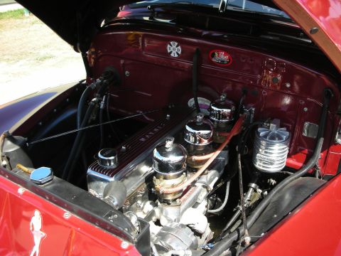 1998 Ford Crown Victoria - Engine bay of a maroon custom car showing a dressed-up flathead-style V8 with finned valve covers, chrome fittings and a Maltese-cross embl…