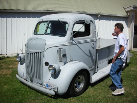 1998 Ford Crown Victoria - primer-gray vintage cab-over-engine pickup with chrome wheels sits on grass by a white building as a man in jeans walks past inspecting it.