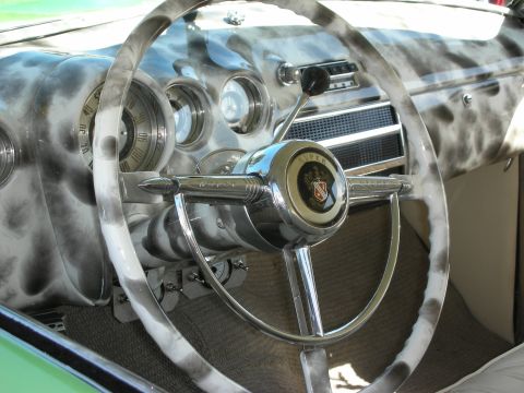 1998 Ford Crown Victoria - Interior detail of a 1950s-era car showing a two-spoke marbled white steering wheel, chrome horn ring and a row of round chrome-bezel gauge…