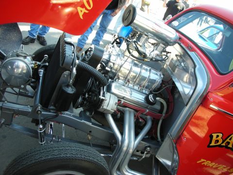 1998 Ford Crown Victoria - Open front bay of a red hot rod showing a polished supercharged V8 with zoomie-style headers and a radiator, spectators standing behind it.