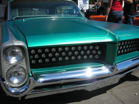 1998 Ford Crown Victoria - Front end of a teal metallic custom car featuring stacked headlights and a black grille studded with chrome bullet ornaments, people behind…