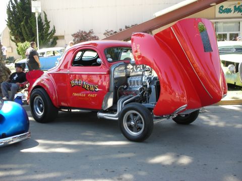 1998 Ford Crown Victoria - red high-stance gasser coupe lettered 'Bad News Travels Fast' with its hood tilted up to reveal a blown engine, parked on a street at a car…