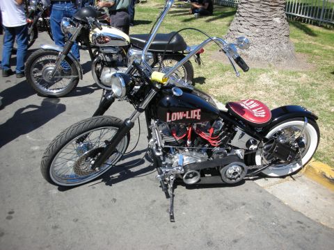 1998 Ford Crown Victoria - black bobber-style chopper with ape-hanger bars, a 'Low-Life' lettered tank and a red-accented V-twin engine on its kickstand at a show.