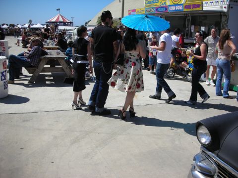 1998 Ford Crown Victoria - group of rockabilly-styled show attendees, one carrying a blue parasol and another in a red floral dress, mingle by food booths on a sunny…