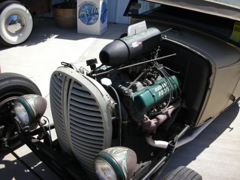 1998 Ford Crown Victoria - Open engine bay of a tan rat-rod roadster showing a green-painted vintage V8 lettered 'Buick Rocket' with a finned grille shell on the pave…