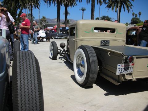 1998 Ford Crown Victoria - Rear three-quarter view of a tan chopped rat-rod pickup with green pinstriping and whitewall wheels at a palm-lined outdoor car show.