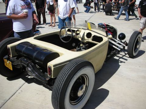 1998 Ford Crown Victoria - low tan open-wheel roadster rat rod with exposed engine, bench seat and steering column sits on pavement surrounded by standing spectators.