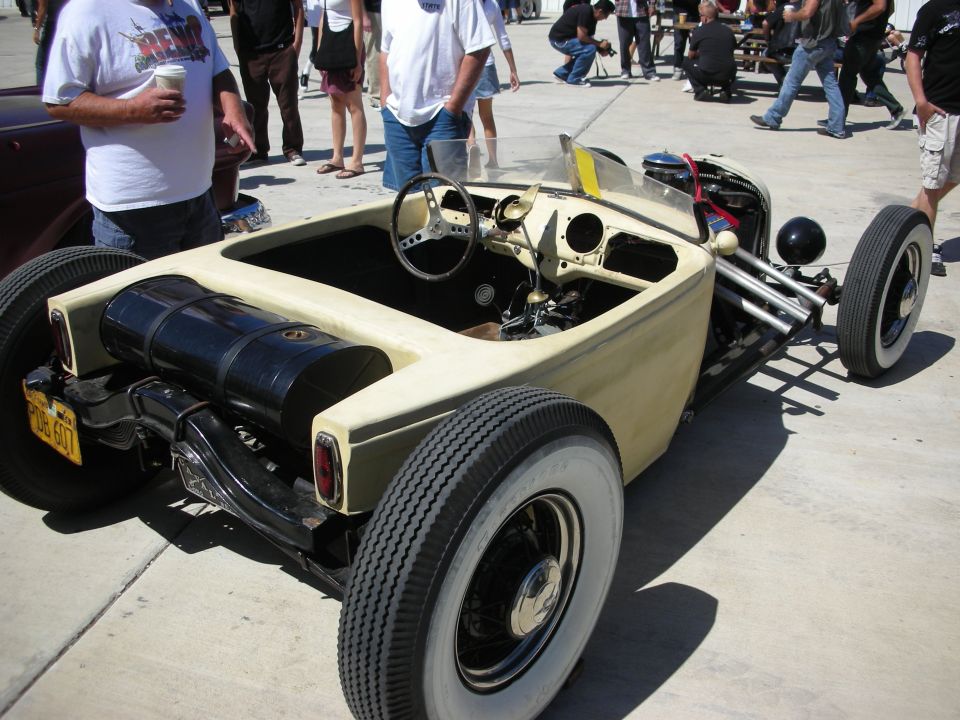 Tan low roadster rat rod with exposed engine and steering, spectators standing around