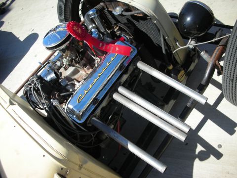 1998 Ford Crown Victoria - Engine bay of a tan rat rod showing a chrome-dressed Cadillac-lettered V8 with a red award ribbon and four polished aluminum exhaust tubes.