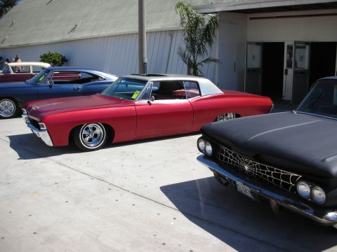 1998 Ford Crown Victoria - matte-red lowered coupe parked between a black classic with spiderweb-painted hood and other vintage cars on pavement.