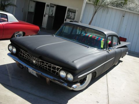 1998 Ford Crown Victoria - flat-black finned late-1950s sedan with spiderweb grille artwork and a serape-draped interior, parked outside a shop.