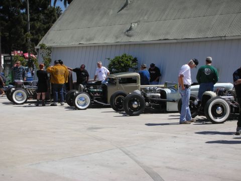 1998 Ford Crown Victoria - row of low-slung rat-rod hot rods with whitewall tires lined up on pavement as spectators mill about near a barn.