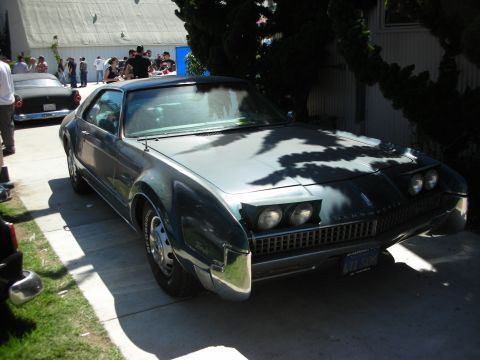 1998 Ford Crown Victoria - teal late-1960s Oldsmobile Toronado coupe with hidden-headlight styling parked on a driveway, a crowd gathered behind it.