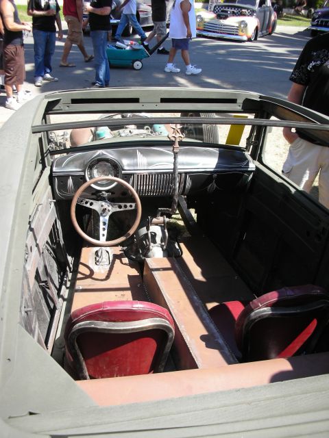1998 Ford Crown Victoria - open cockpit of a topless hot rod showing a banjo steering wheel, painted dash and weathered bench seats over a bare floor.
