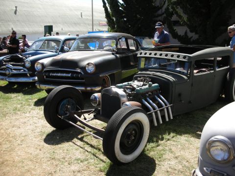 1998 Ford Crown Victoria - flat-black chopped rat rod with an exposed V8 and zoomie side pipes parked on grass beside a black classic sedan.