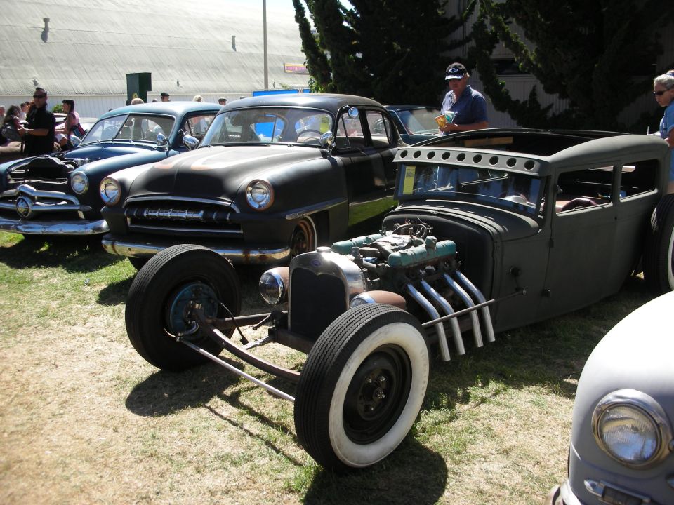 Matte black chopped rat rod with exposed V8 and side pipes beside black classic