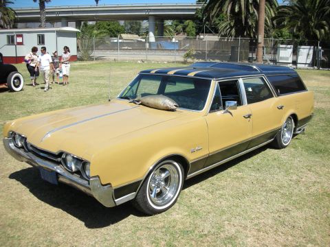 1998 Ford Crown Victoria - gold late-1960s Oldsmobile station wagon with a black roof and chrome wheels parked on grass under a freeway overpass.