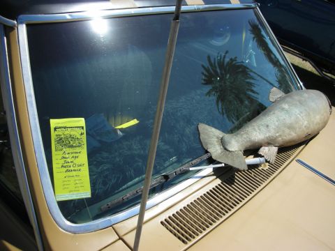 1998 Ford Crown Victoria - gray fish-shaped sculpture rests on the cowl of a gold car, with a handwritten show entry card tucked under the windshield.