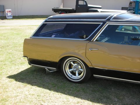 1998 Ford Crown Victoria - rear quarter of a gold station wagon with black roof, chrome wheels and whitewalls, a vintage pickup parked behind.