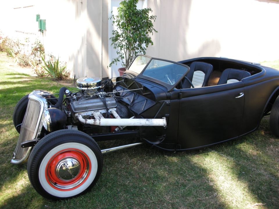 Black 1930s roadster hot rod with exposed V8, whitewall tires, and chrome wheels on lawn