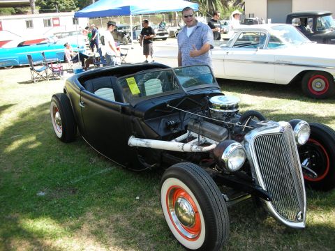 1998 Ford Crown Victoria - satin-black highboy roadster hot rod with exposed V8 and whitewall tires is displayed at a car show as a man gives a thumbs up beside it.