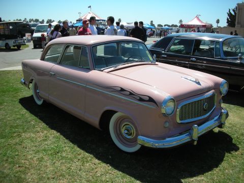1998 Ford Crown Victoria - pink late-1950s two-door sedan with black flame graphics and wide whitewall tires sits on grass at a crowded car show.