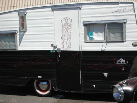 1998 Ford Crown Victoria - Side of a lowered vintage travel trailer in black and white with red pinstriping, a tiki-style face panel, and whitewall tire.
