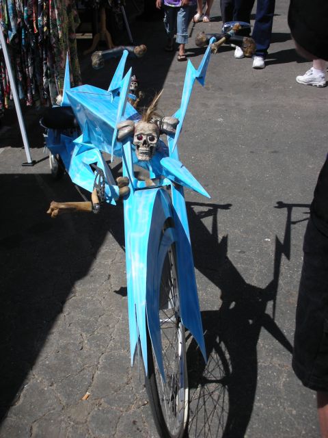 1998 Ford Crown Victoria - Front view of a blue custom art bicycle topped with a horned skull and bone accents, standing on pavement among onlookers.