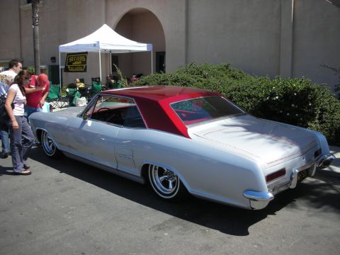 1998 Ford Crown Victoria - silver 1960s personal-luxury hardtop coupe with a red vinyl roof and wire wheels parked near a vendor tent at a car show.