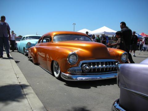 1998 Ford Crown Victoria - copper-orange chopped and lowered 1950s lead-sled custom coupe with whitewall tires sits among other customs at a car show.