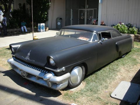 1998 Ford Crown Victoria - flat-black chopped and lowered 1950s-style custom car with pinstriping sits on grass beside a building at a car show.