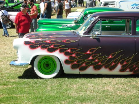 1998 Ford Crown Victoria - lowered 1950s sedan painted purple and white with yellow-edged flames and green wheels at a crowded car show.
