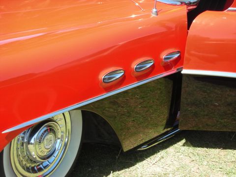 1998 Ford Crown Victoria - Close-up of an orange custom car body panel with three chrome oval portholes above a chrome hubcap and whitewall.