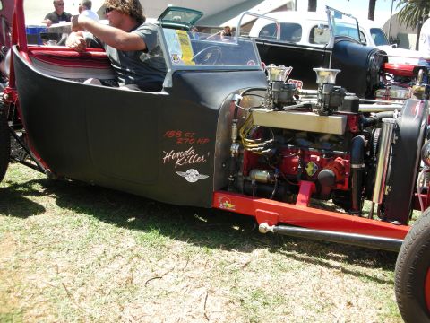 1998 Ford Crown Victoria - man sits in a black T-bucket hot rod with an exposed inline engine and Honda Killer lettering at a car show.