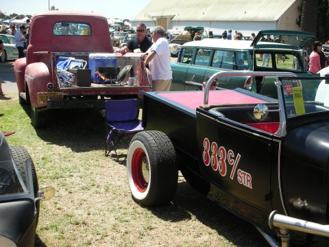 1998 Ford Crown Victoria - black open roadster lettered 333 c/STR with red bed sits beside a maroon vintage pickup at a crowded car show.