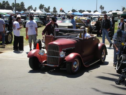 1998 Ford Crown Victoria - maroon primer-finish open roadster surrounded by spectators on the asphalt at a busy outdoor car show.