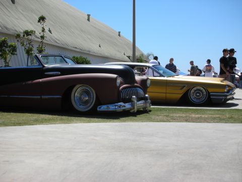 1998 Ford Crown Victoria - maroon-primer chopped convertible and a gold-painted hardtop sit lowered side by side at an outdoor car show with spectators behind.