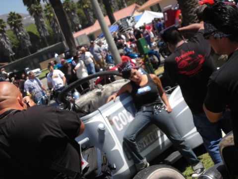 1998 Ford Crown Victoria - busy outdoor car-and-bike show with a woman in a black tank top leaning against a gray lettered hot rod amid the crowd.