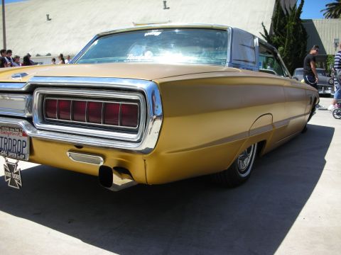 1998 Ford Crown Victoria - Rear quarter of a gold mid-1960s Ford Thunderbird hardtop with vertical taillights and a chrome cross emblem on the license plate frame.