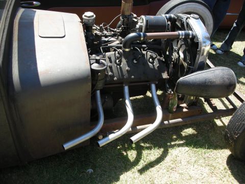 1998 Ford Crown Victoria - rat rod's exposed engine with stacked exhaust pipes mounted in a bare frame, photographed from the side on a grass lot.