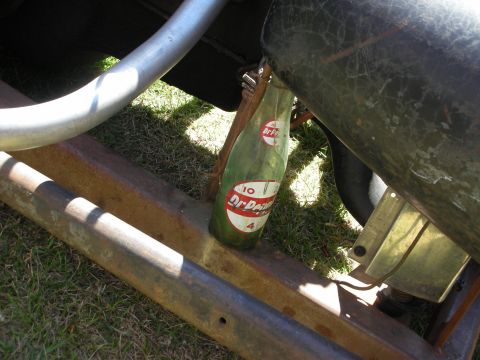 1998 Ford Crown Victoria - old green Dr Pepper glass bottle sits propped on the rusty frame rail of a hot rod over grass in sunlight.